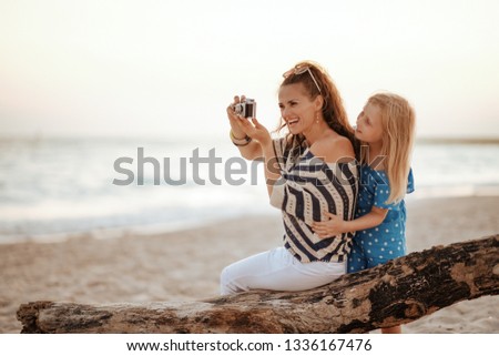 smiling young mother and daughter tourists sitting on a wooden snag and taking photos with retro photo camera on the seacoast in the evening.