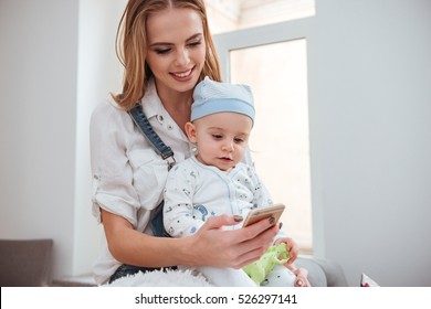 Smiling Young Mother Holding Her Little Son And Using Cell Phone At Home