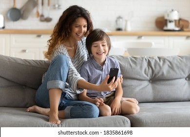 Smiling Young Mother And Little Son Using Phone Together, Hugging, Sitting On Cozy Couch At Home, Happy Mum With Adorable Kid Boy Taking Selfie, Shooting Vlog, Enjoying Leisure Time With Gadget