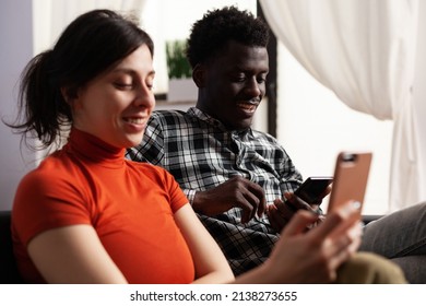 Smiling young multiethnic couple using smartphones while browsing on internet and watching online funny videos. People spending time on phones without interacting to each other.