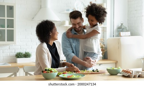 Smiling Young Multiracial Mom And Dad Cook Healthy Delicious Vegetarian Salad With Ethnic Small Daughter. Happy Multiethnic Family With Girl Child Have Fun Prepare Food In Kitchen. Adoption Concept.