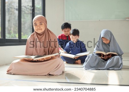 Smiling young Muslim Asian teen girl looking at camera while sitting on mosque floor after reading Quran with her friends on the background