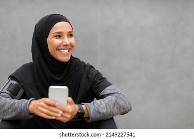 Smiling Young Muslim Woman Athlete In Hijab Sportswear With Smartphone Looking At Empty Space On Gray Wall Background. Relax After Training, Tech For Workout, Sports In City, Body Care Due Covid-19