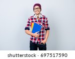 Smiling young nerdy stylish student hipster is standing with books on pure background in black trendy glasses and hat, casual bright checkered outfit