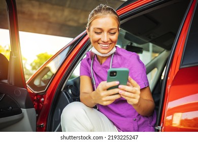 A Smiling Young Nurse Dressed In Her Scrubs Uniform Sitting In Her Car Holding Her Mobile Phone, Taking A Break From Work.