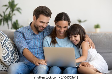 Smiling young parents have fun rest on sofa watching funny video on laptop with little girl child, happy family mom and dad relax on couch with preschooler daughter enjoy movie laughing at home