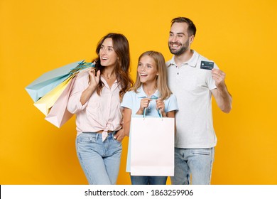 Smiling Young Parents Mom Dad With Child Kid Daughter Teen Girl In Basic T-shirts Hold Package Bags With Purchases Credit Bank Card Isolated On Yellow Background Studio Portrait. Family Day Concept