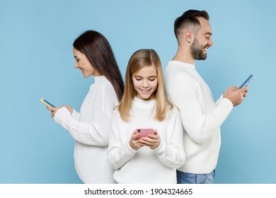 Smiling Young Parents Mom Dad With Child Kid Daughter Teen Girl In White Sweaters Using Mobile Phone Typing Sms Message Isolated On Blue Background Studio. Family Day Parenthood Childhood Concept