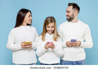 Smiling Young Parents Mom Dad With Child Kid Daughter Teen Girl In White Sweaters Using Mobile Phone Typing Sms Message Isolated On Blue Background Studio. Family Day Parenthood Childhood Concept