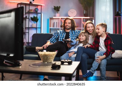 Smiling Young Parents With Two Kids Sitting On Couch And Watching TV. Happy Caucasian Family In Casual Wear Spending Evening Time Together At Home.