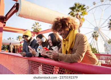 Smiling young persons using cell phones. Teenagers addicted to online technological trends. Group of friends leaning on a railing checking their social networks. People gathered at the amusement park.