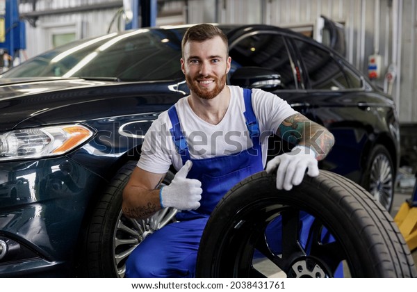 Smiling young positive professional technician\
car mechanic man wears denim blue overalls white t-shirt stand hold\
wheel show thumb up gesture work in light modern vehicle repair\
shop workshop indoors