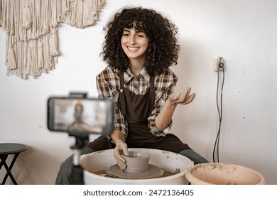 Smiling young potter woman sculpting a vase on potter's wheel while recording a video blog using a smartphone. - Powered by Shutterstock