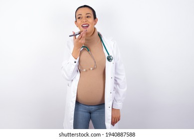 Smiling young pregnant doctor woman wearing medical uniform against white background sending voice message on her smart phone. Communication and new technologies concept.