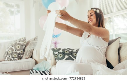 Smiling Young Pregnant Woman Sitting On Sofa With Presents At Baby Shower Party. Expecting Mother Looking At Clothes Of Child And Smiling.