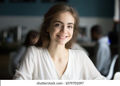 Smiling young pretty lady looking at camera in public place, adolescent girl with cute pretty face posing indoors, happy millennial casual woman student or professional in cafe head shot portrait