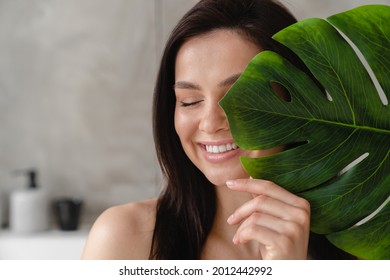 Smiling Young Pretty Woman Hiding Her Face With Tropical Leaf In Bathroom. Spa Procedures At Home Beautician. Skin And Hair Care Treatment.