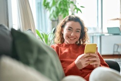 Smiling Young Pretty Woman Sitting On Couch Using Apps On Cell Phone Technology, Happy Lady Holding Smartphone In Hands, Looking At Camera, Relaxing On Sofa With Cellphone Checking Cellular Device.