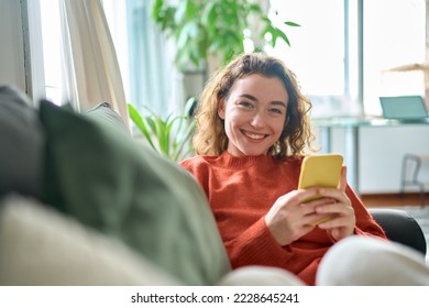 Smiling young pretty woman sitting on couch using apps on cell phone technology, happy lady holding smartphone in hands, looking at camera, relaxing on sofa with cellphone checking cellular device.