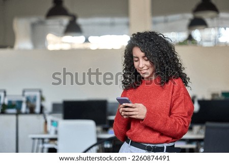 Smiling young professional latin business woman, happy lady corporate leader holding cellular phone working standing in modern office using mobile apps cellphone technology device looking at cell. 商業照片 © 