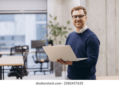 Smiling young professional man using a laptop in a modern office setting, showcasing a tech savvy and productive workplace atmosphere