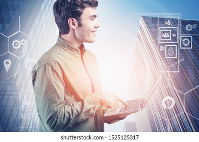 Smiling young programmer wearing green shirt using laptop standing over cityscape background with double exposure of HUD digital interface. Concept of hi tech in business and education. Toned image