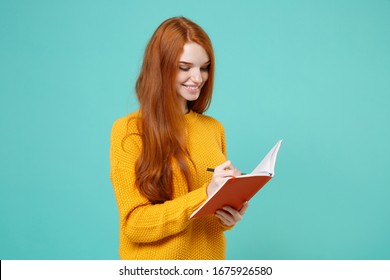 Smiling Young Redhead Woman Girl In Yellow Knitted Sweater Posing Isolated On Blue Turquoise Wall Background Studio Portrait. People Lifestyle Concept. Mock Up Copy Space. Writing Notes In Notebook