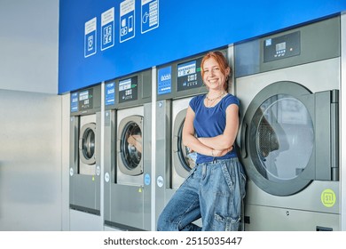 Smiling young redhead woman standing in a public laundromat looking at camera with washing machines in the background - Powered by Shutterstock