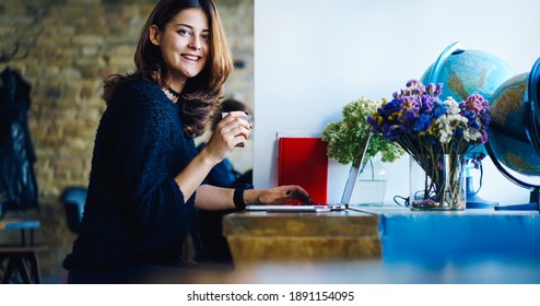 Smiling Young Remote Worker Sitting In Cafe While Working On Laptop And Drinking Coffee With Flower And Globe At Table And Looking At Camera