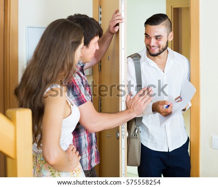 Smiling young salesman came home to the serious merried couple and stands in doorway