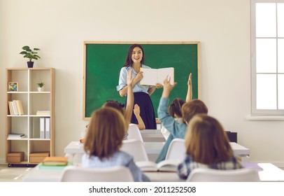 Smiling Young School Teacher Standing In Front Of Blackboard In Classroom And Showing Mockup Book To Group Of Elementary Students Who Are Raising Their Hands Up. Education Concept