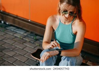 Smiling young skater girl with skate-board sitting in sunset light. Blonde woman using smart watch on red background. Summer outdoor activity, concept of freedom, happiness and healthy lifestyle.