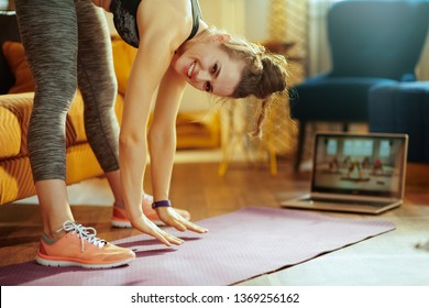 Smiling Young Sports Woman In Sport Clothes At Modern Home Using Laptop To Watch Fitness Streaming On Internet While Doing Stretching On Fitness Mat.