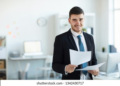 Smiling young successful banker in elegant suit looking through financial documents in office
