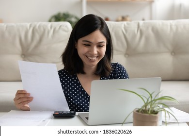 Smiling young Vietnamese woman look at laptop screen paying bills taxes online use easy banking system. Happy asian female manage budget at home, consider household finances expenditures on computer.