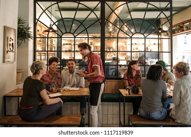 Smiling young waiter taking orders from a diverse group of customers sitting together at a restaurant table - Powered by Shutterstock