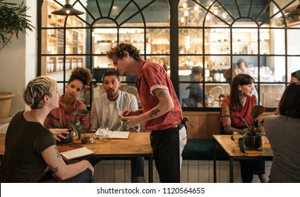 Smiling young waiter taking orders from a group of customers sitting together at a table in a bistro - Powered by Shutterstock