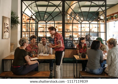 Smiling young waiter taking orders from a diverse group of customers sitting together at a restaurant table