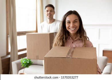 Smiling young wife holding cardboard box relocating into new home with husband looking at camera packing, happy couple first time real estate buyers renters owners tenants buy rent house concept