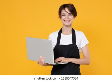 Smiling Young Woman 20s Barista Bartender Barman Or Grocery Employee In White T-shirt Apron Posing Working On Laptop Pc Computer Looking Camera Isolated On Yellow Color Wall Background Studio Portrait