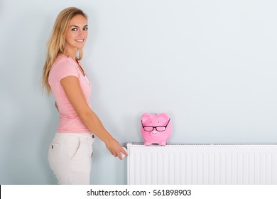 Smiling Young Woman Adjusting Thermostat To Reduce Heating To Save Money On Energy Bill