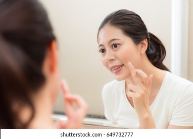 smiling young woman applying face cream on finger ready smear face and looking at mirror in bathroom showing beauty skin care.