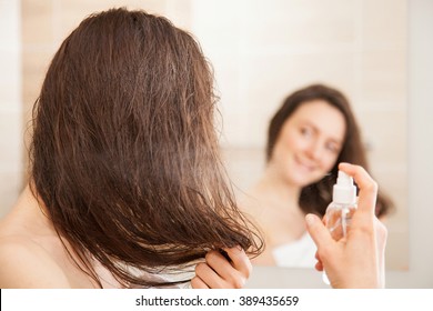 Lächelnde junge Frau, die vor einem Spiegel sprüht; Haarpflegekonzept