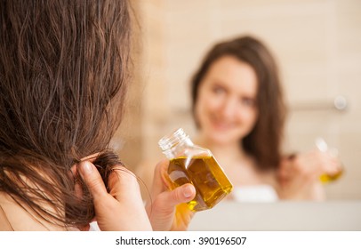 Smiling young woman applying oil mask to hair tips in front of a mirror; haircare concept