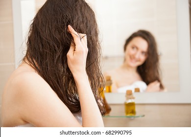 Smiling Young Woman Applying Oil Mask To Her Hair In Front Of A Mirror; Haircare Concept