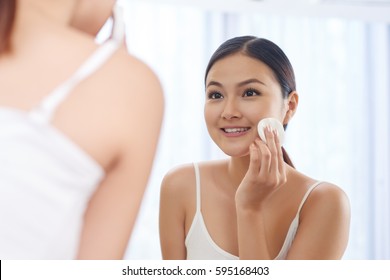 Smiling Young Woman Applying Toner On Her Face In Front Of Mirror