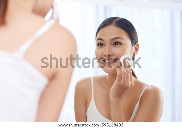 Smiling young woman applying toner on her face in
front of mirror