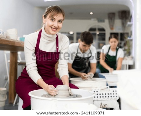 Smiling young woman in apron modeling clay dish from row material using pottery wheel in workroom. Persons making pottery products