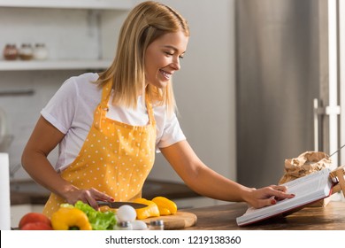 Smiling Young Woman In Apron Reading Cookbook While Cooking In Kitchen