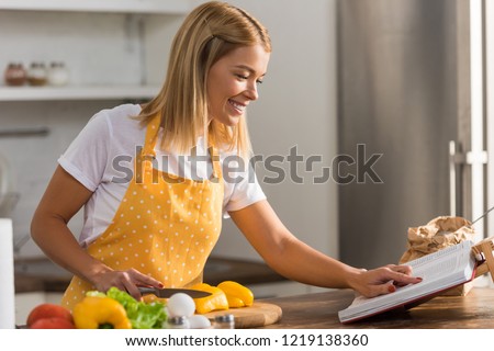 smiling young woman in apron reading cookbook while cooking in kitchen